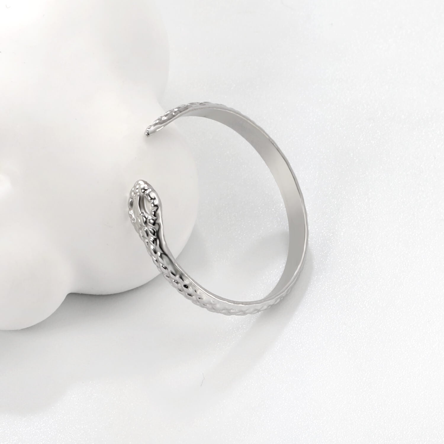 Madoww brändin Cobra vedenkestävä ruostumattomasta teräksestä valmistettu hopean värinen sormus. Sormuksessa on kobran pää ja se nojaa valkoista elementtiä vasten.