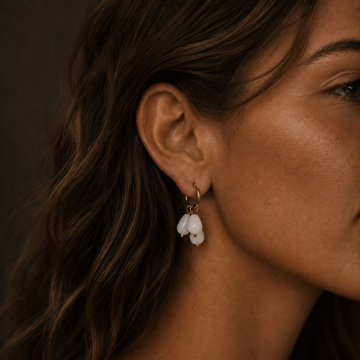 Close-up of a woman wearing gold hoop earrings with three frosted white teardrop stones, photographed in warm soft lighting with the jewelry as the main focus.