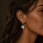 Close-up of a woman wearing gold hoop earrings with three frosted white teardrop stones, photographed in warm soft lighting with the jewelry as the main focus.
