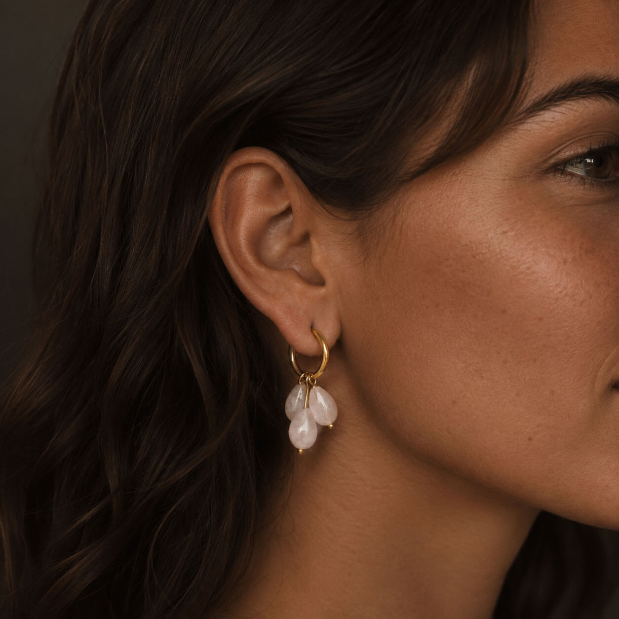 Close-up of a woman wearing gold hoop earrings with three soft rose-colored teardrop stones, photographed in warm soft lighting with the delicate jewelry as the focal point.