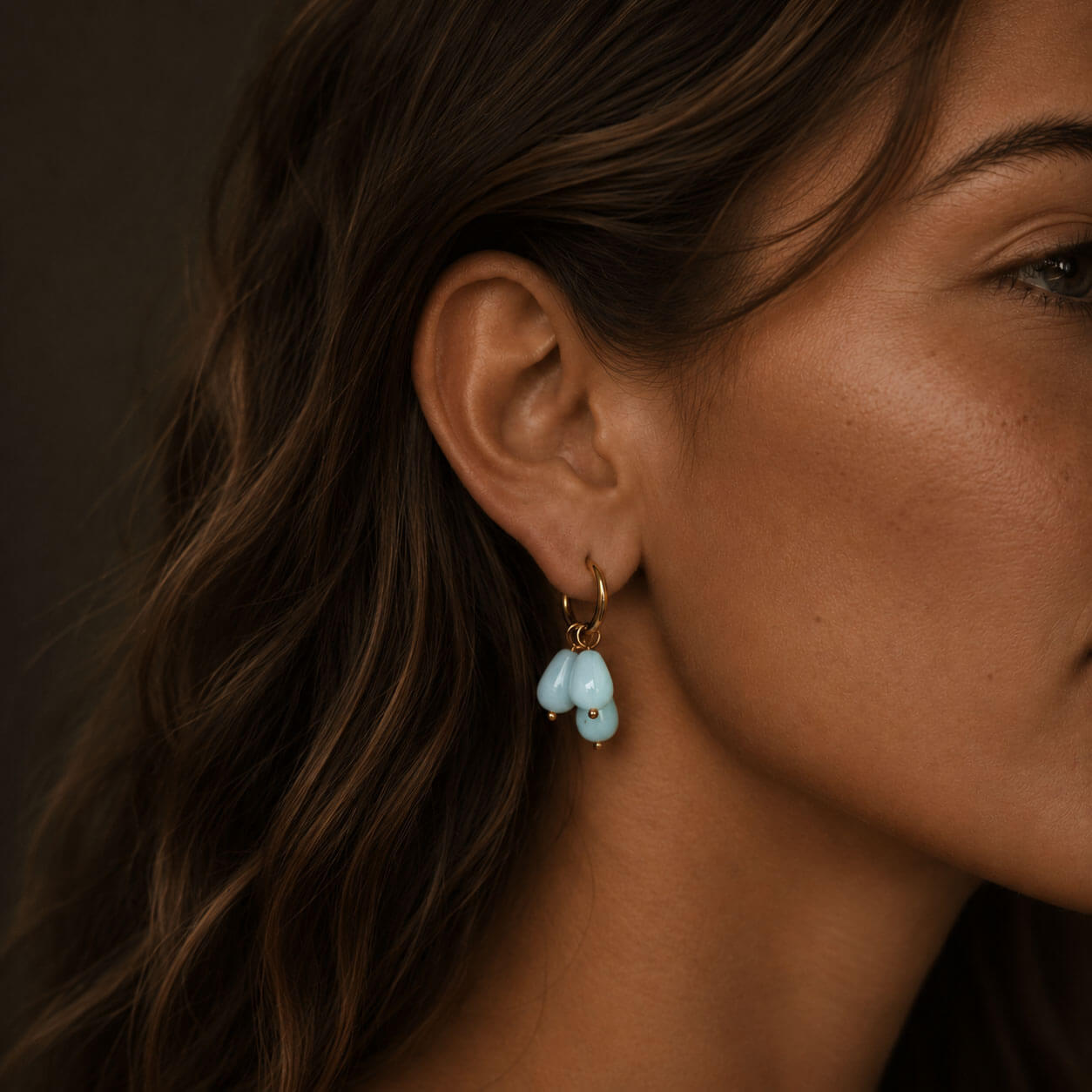 Close-up of a woman wearing gold hoop earrings with three pale blue teardrop stones, photographed in warm soft lighting with the earrings clearly highlighted.