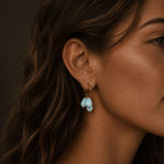 Close-up of a woman wearing gold hoop earrings with three pale blue teardrop stones, photographed in warm soft lighting with the earrings clearly highlighted.