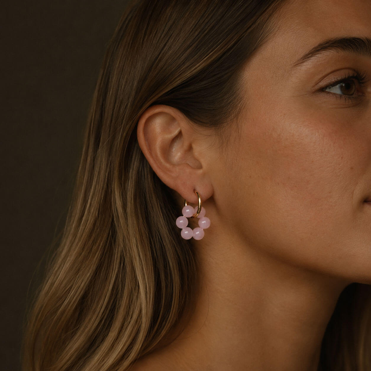 Close-up portrait of a woman wearing gold hoop earrings with soft rose pink stone beads, highlighted as the focal point in a warm, natural side-profile image.