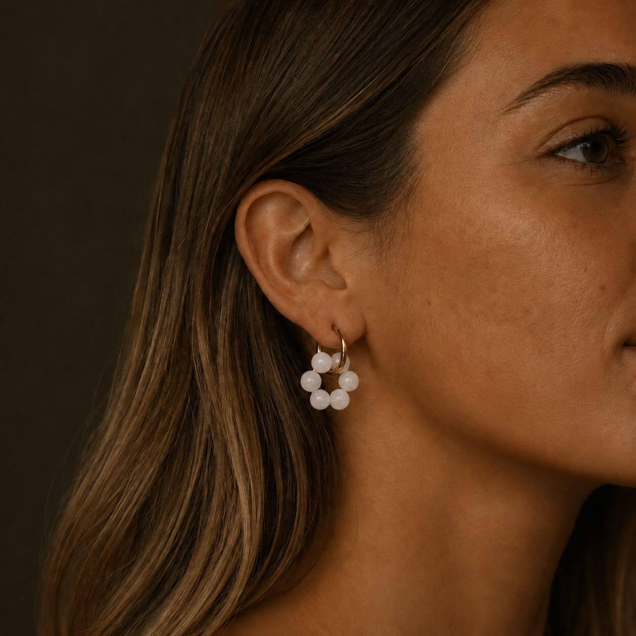 Close-up portrait of a woman wearing gold hoop earrings with translucent milky white stone beads, softly lit against a dark neutral background.