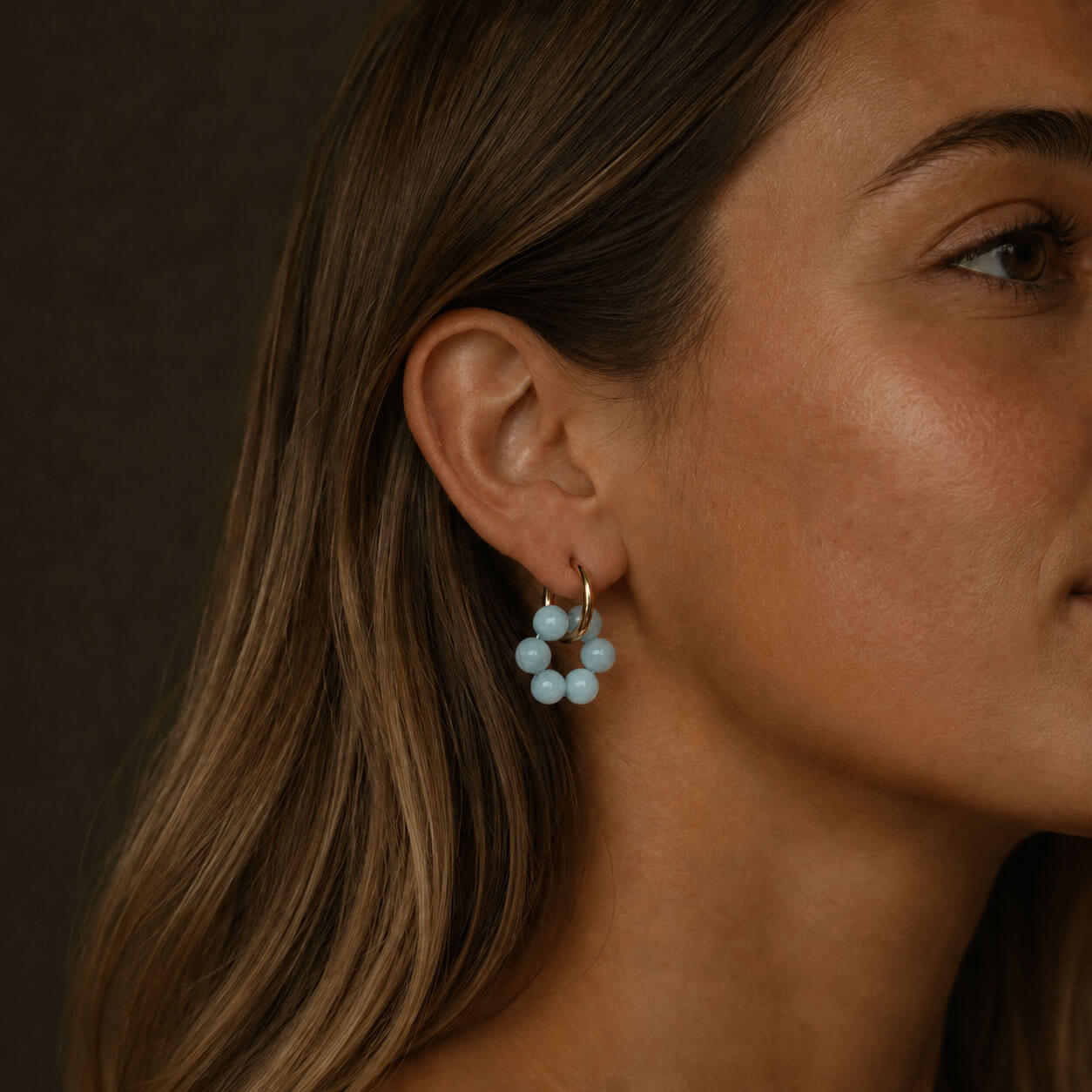 Close-up portrait of a woman wearing gold hoop earrings with pale blue stone beads, the jewelry standing out in warm soft lighting against a dark background.