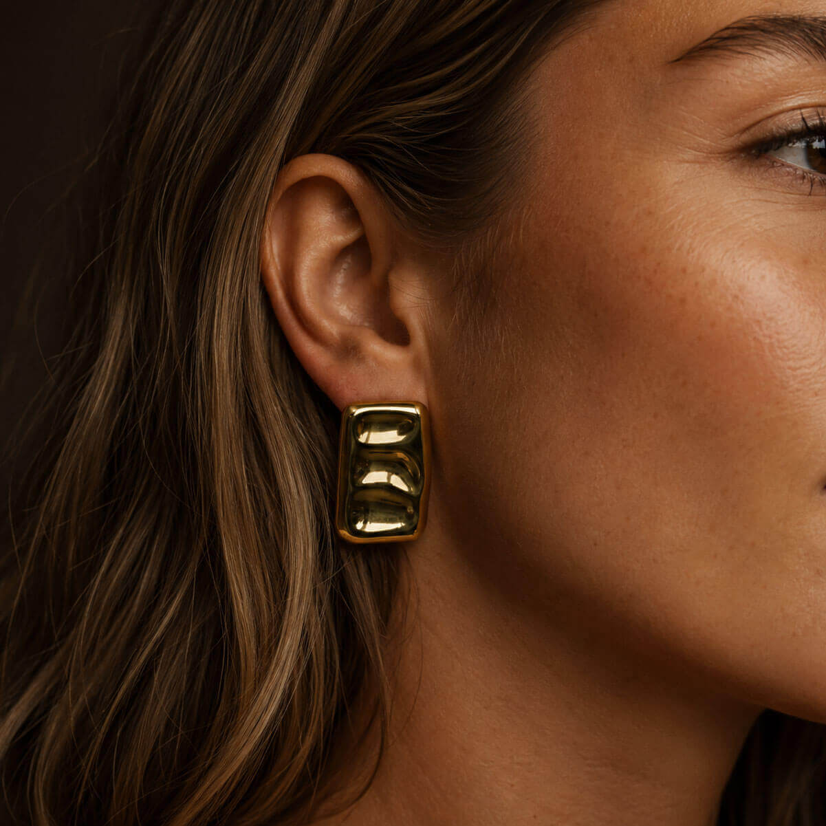 Close-up portrait of a woman wearing bold PVD gold-plated block earrings, with the sculptural jewelry highlighted against warm dark lighting.