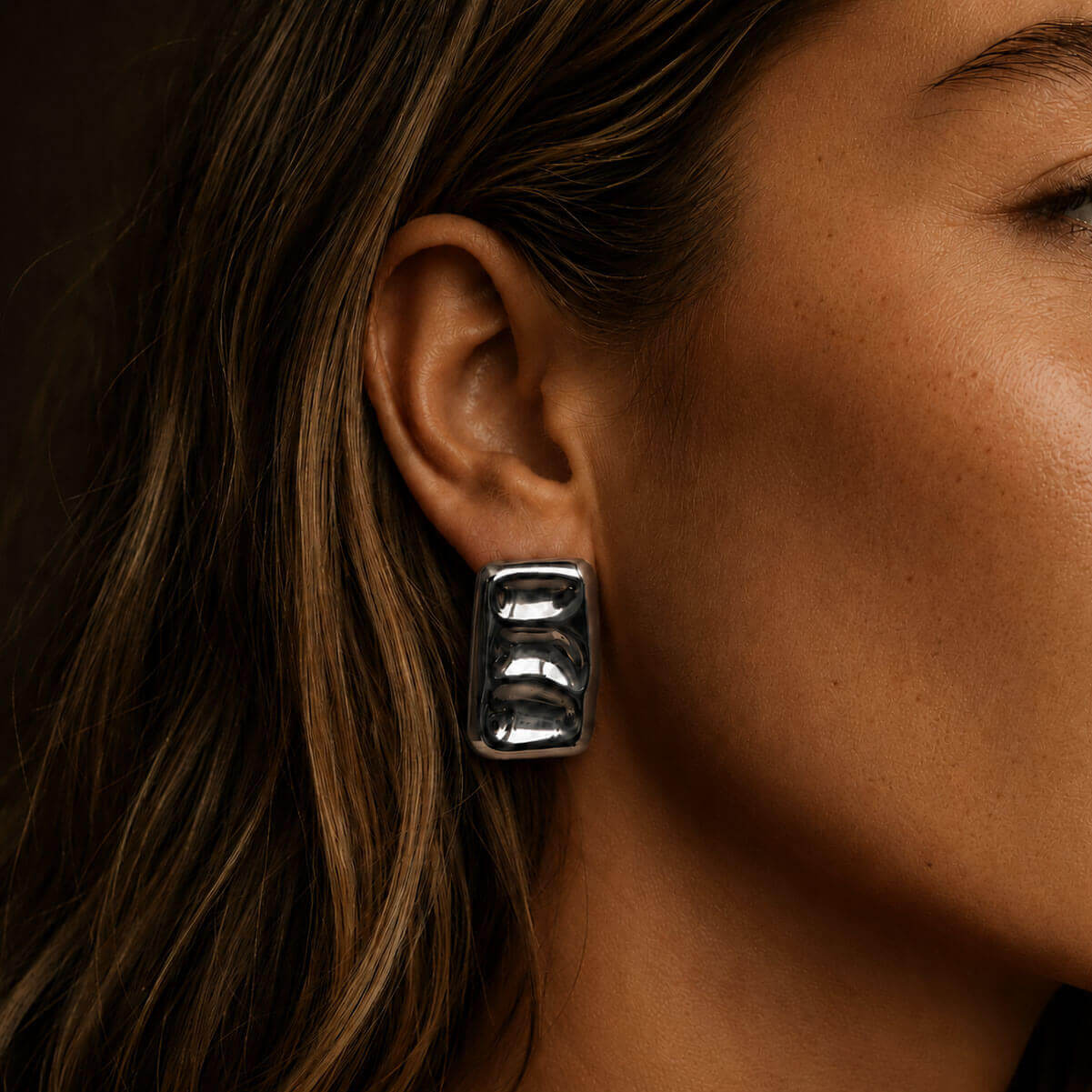 Close-up portrait of a woman wearing bold silver-colored block earrings, with the reflective sculptural jewelry as the main focus against a dark background.