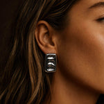 Close-up portrait of a woman wearing bold silver-colored block earrings, with the reflective sculptural jewelry as the main focus against a dark background.