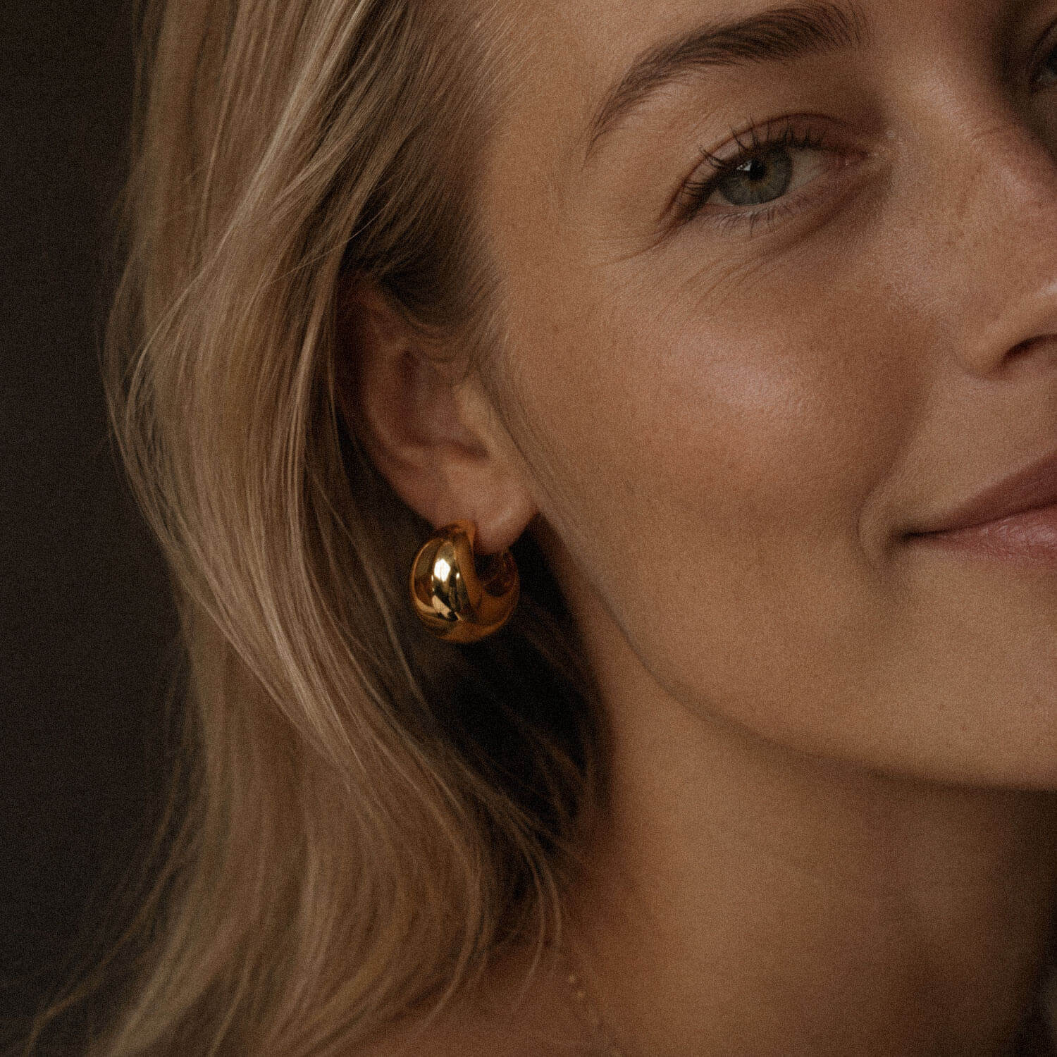 Close-up beauty portrait of a blonde woman with glowing skin wearing a chunky gold hoop earring made from 18k PVD gold-plated stainless steel.