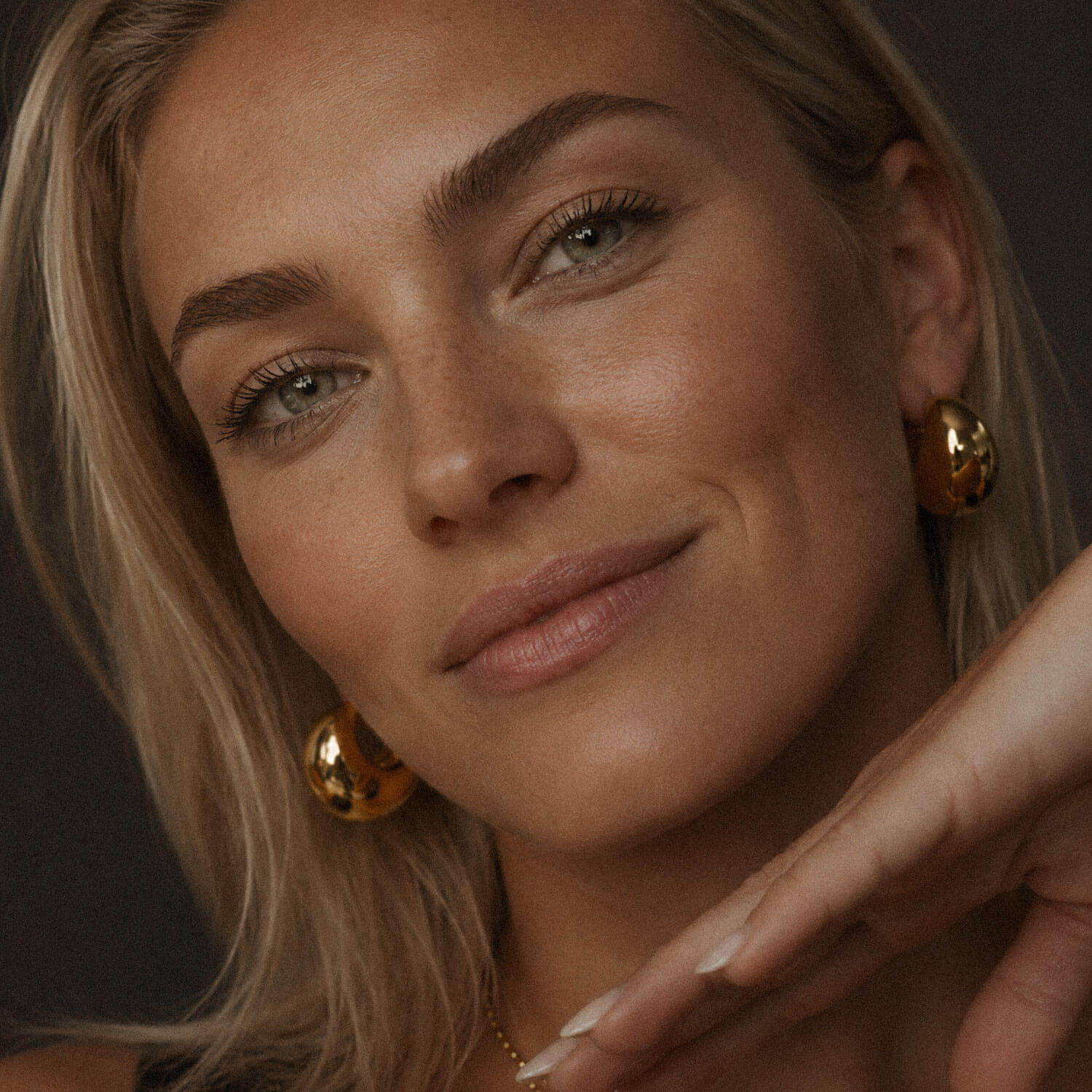 Softly lit close-up portrait of a blonde woman wearing chunky gold hoop earrings made from 18k PVD gold-plated stainless steel, with natural makeup and a delicate necklace.