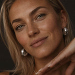 Soft studio portrait of a blonde woman wearing chunky silver-tone hoop earrings made from stainless steel, with natural makeup, glowing skin, and a delicate necklace.