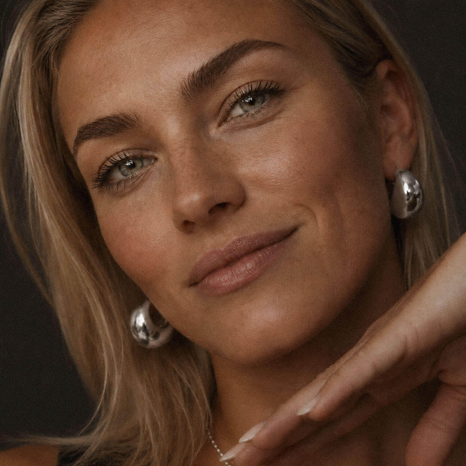 Soft studio portrait of a blonde woman wearing chunky silver-tone hoop earrings made from stainless steel, with natural makeup, glowing skin, and a delicate necklace.