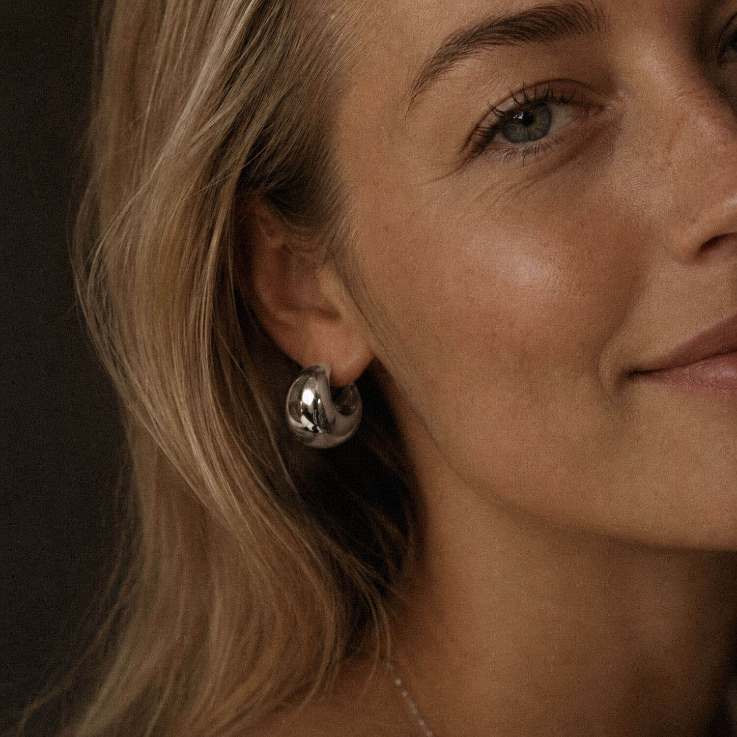 Close-up beauty portrait of a blonde woman with luminous skin wearing a chunky silver-tone hoop earring made from stainless steel, edited to match the same photoshoot style.