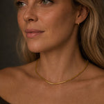 Close-up portrait of a woman wearing a delicate 18k PVD gold-plated stainless steel snake chain necklace. The necklace rests naturally along the collarbone, with soft warm lighting and a dark neutral background highlighting the jewelry.
