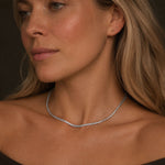 Close-up portrait of a woman wearing a delicate silver-tone stainless steel snake chain necklace. The necklace rests naturally along the collarbone, with soft warm lighting and a dark neutral background emphasizing the refined jewelry design.