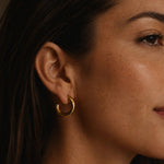 Close-up side profile of a woman wearing gold hoop earrings made from stainless steel with 18K PVD gold plating, shown in warm soft lighting against a dark neutral background.