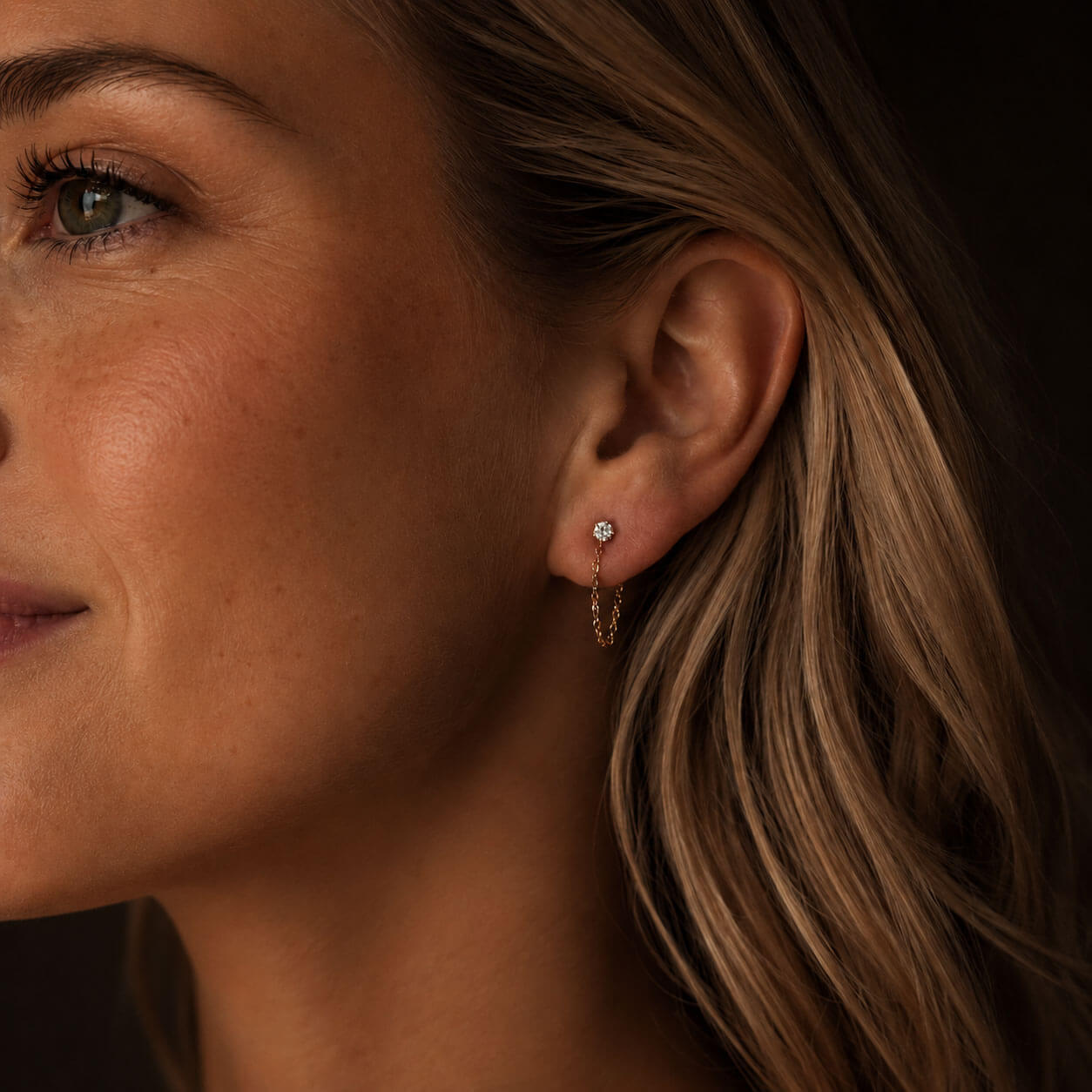 Warm, moody close-up side portrait of a Scandinavian-looking woman wearing delicate 18K PVD gold-plated stainless steel chain earrings with zirconia details. The dark background and soft lighting highlight the small sparkling stud and fine looped chain.