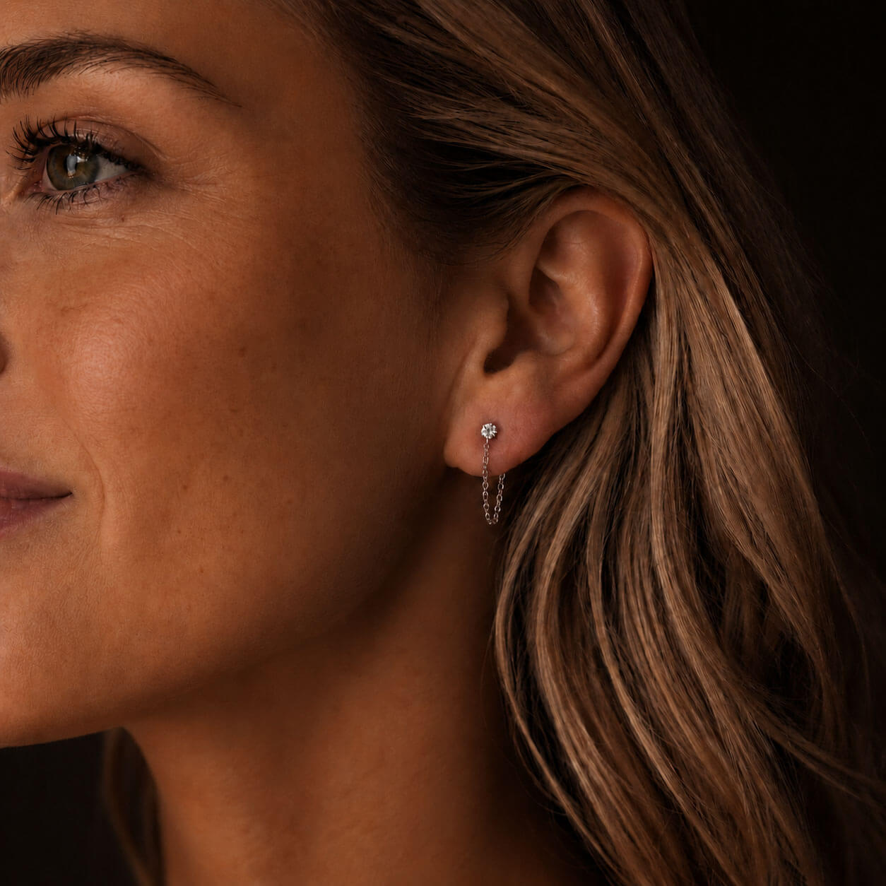 Warm, moody close-up side portrait of a Scandinavian-looking woman wearing delicate silver-toned stainless steel chain earrings with zirconia details. The dark background and soft lighting highlight the small sparkling zirconia stud and fine looped chain.
