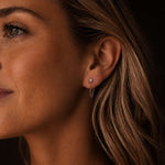 Warm, moody close-up side portrait of a Scandinavian-looking woman wearing delicate silver-toned stainless steel chain earrings with zirconia details. The dark background and soft lighting highlight the small sparkling zirconia stud and fine looped chain.
