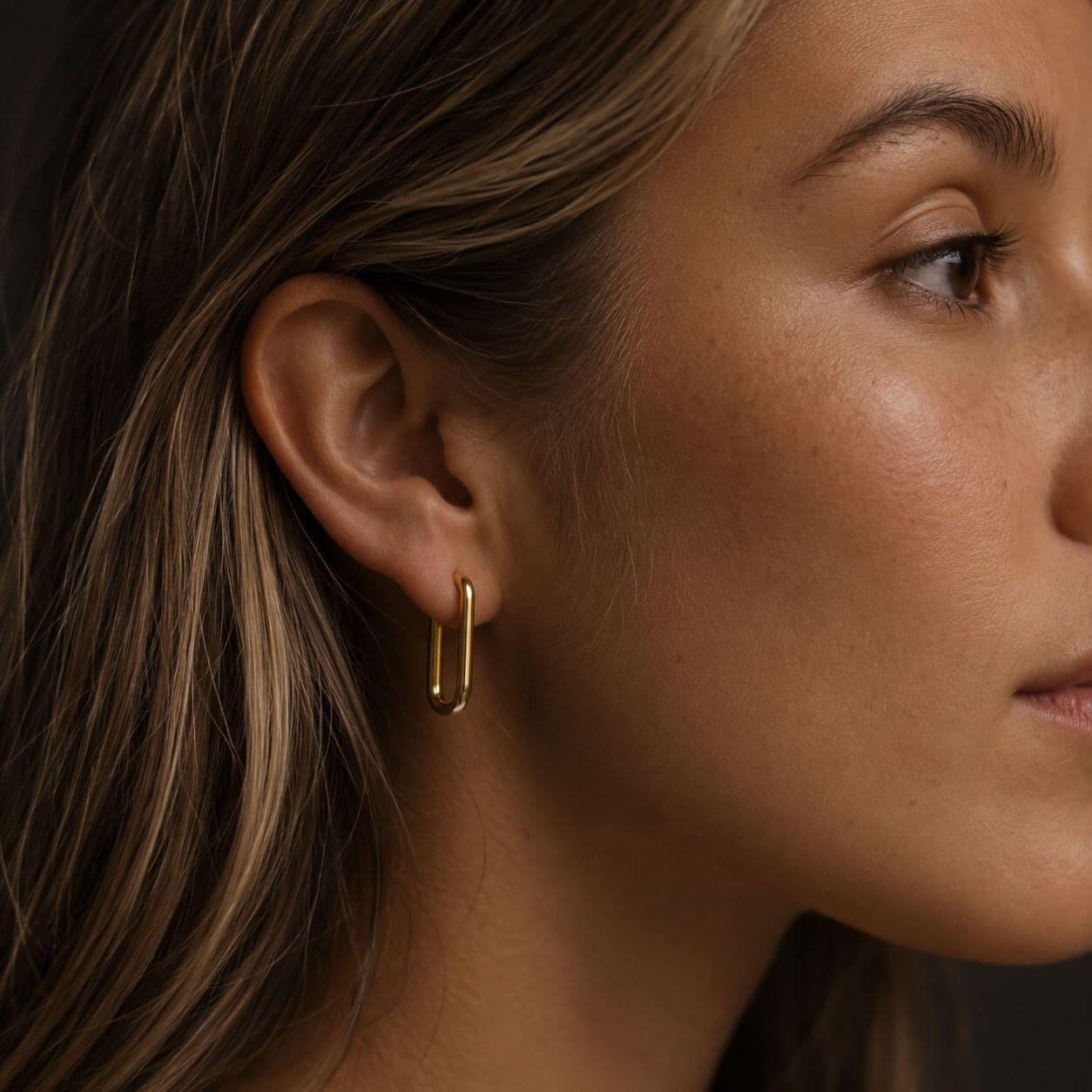 Close-up side profile of a woman wearing a polished gold hoop earring made from 18k PVD gold-plated stainless steel, styled against a dark neutral background with soft warm lighting.