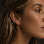 Close-up side profile of a woman wearing a polished gold hoop earring made from 18k PVD gold-plated stainless steel, styled against a dark neutral background with soft warm lighting.