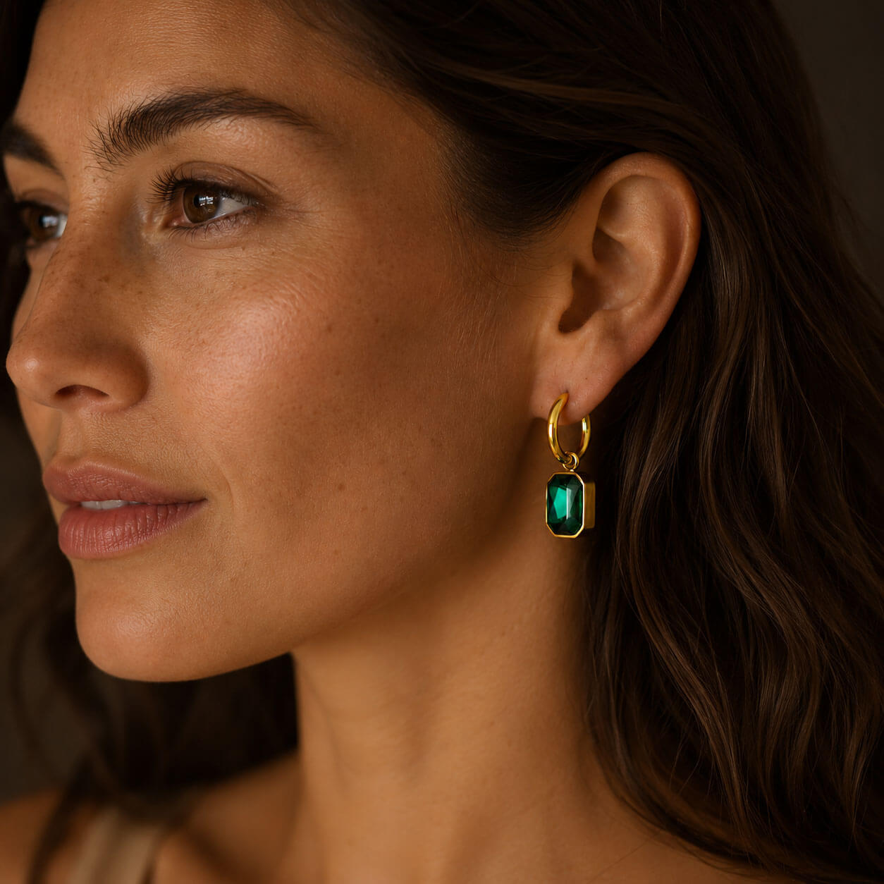 Three-quarter close-up portrait of a woman wearing an 18k PVD gold-plated stainless steel hoop earring with a large green zircon pendant.