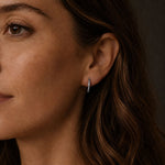 Close-up of a woman with olive-toned skin and freckles wearing a sleek stainless steel hoop earring with zirconia stones, shown from the side in soft, warm lighting against a dark neutral background.