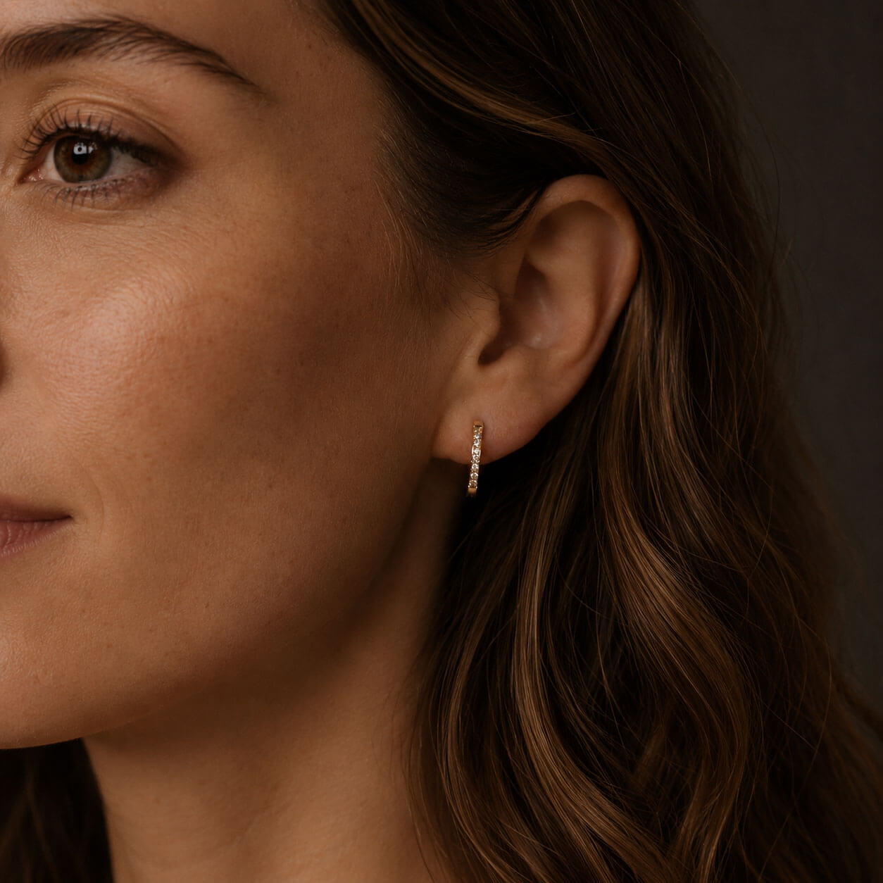 Close-up of a woman with olive-toned skin and freckles wearing a small 18K PVD gold-plated stainless steel hoop earring with sparkling zirconia stones, shown from the side against a warm, dark background.