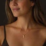 A woman wearing a delicate PVD 18K gold-plated bar necklace in warm, soft lighting. The minimalist gold pendant with madoww logo engraved and fine chain are the focal point against a calm, dark neutral background.