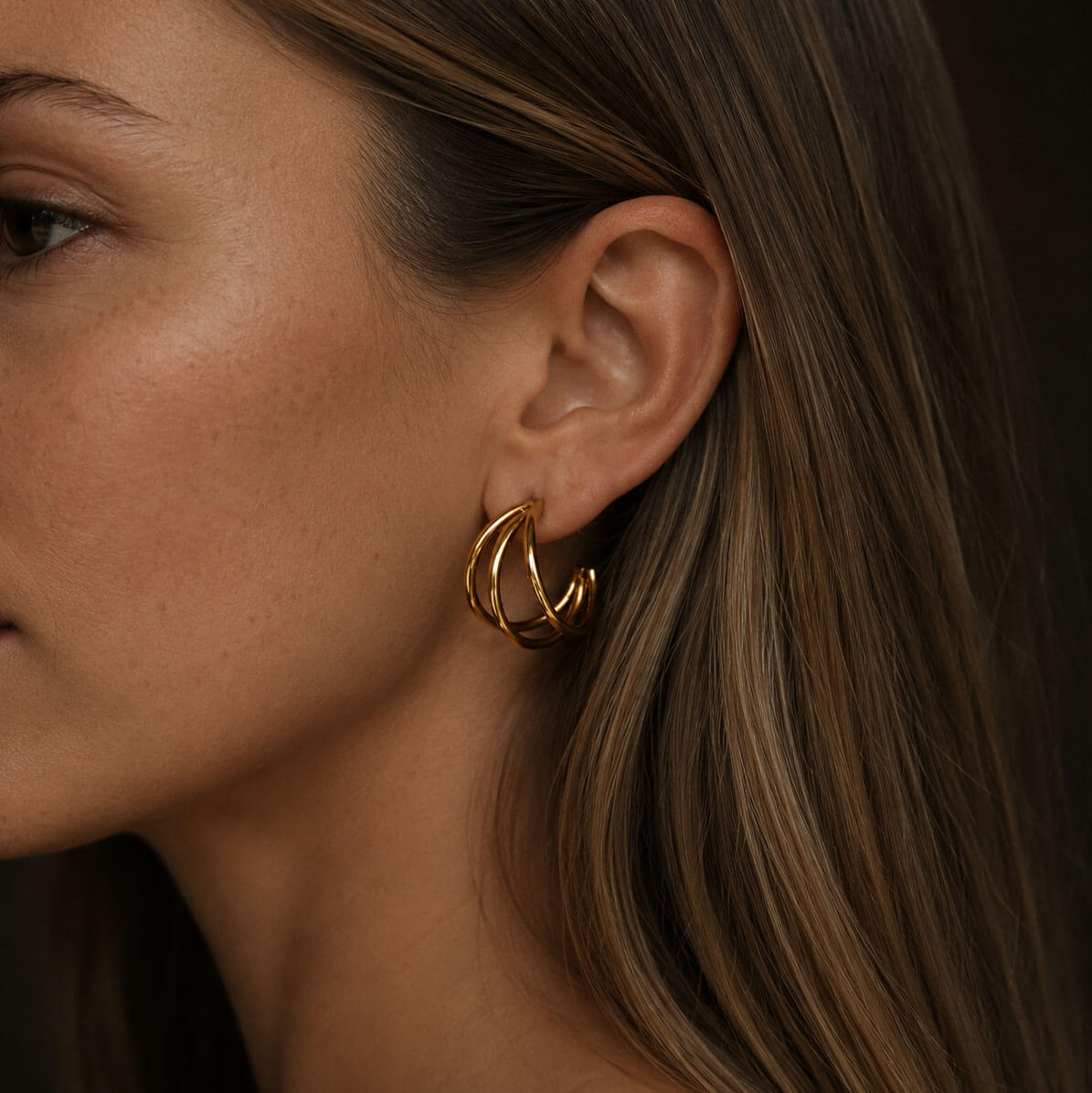 Close-up side profile of a woman wearing the Triple Hoop earring, crafted from stainless steel with 18K PVD gold plating, highlighted against a warm, dark background.